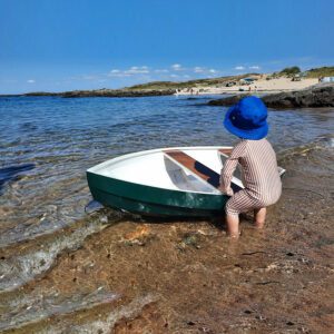 Enfant avec un chapeau bleu jouant avec un petit bateau "Fripouille" sur une plage.