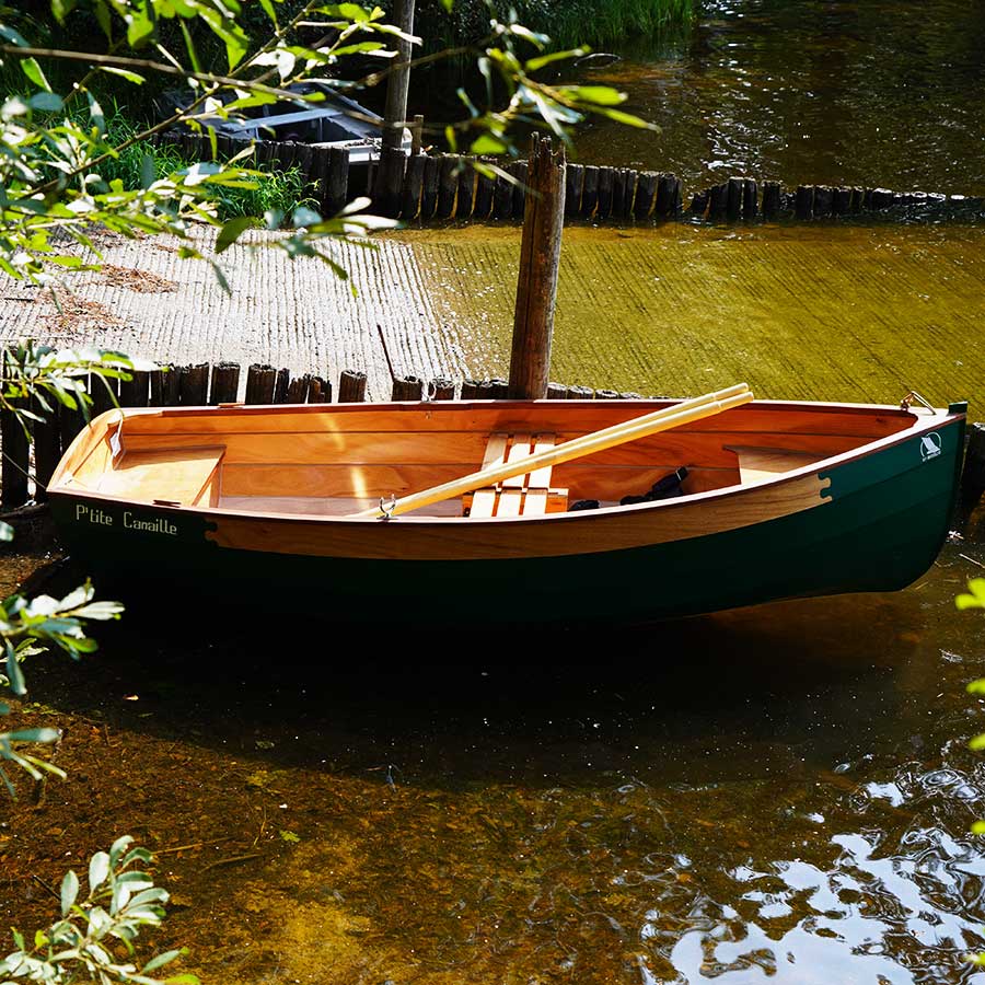 Voilier en bois P’tite Canaille amarré près d’un ponton en bord de lac, sous les arbres.