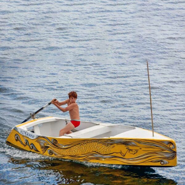 Enfant en maillot rouge naviguant sur un petit bateau de godille en kit, décoré artistiquement, modèle Triton 4.8, sur eau calme.