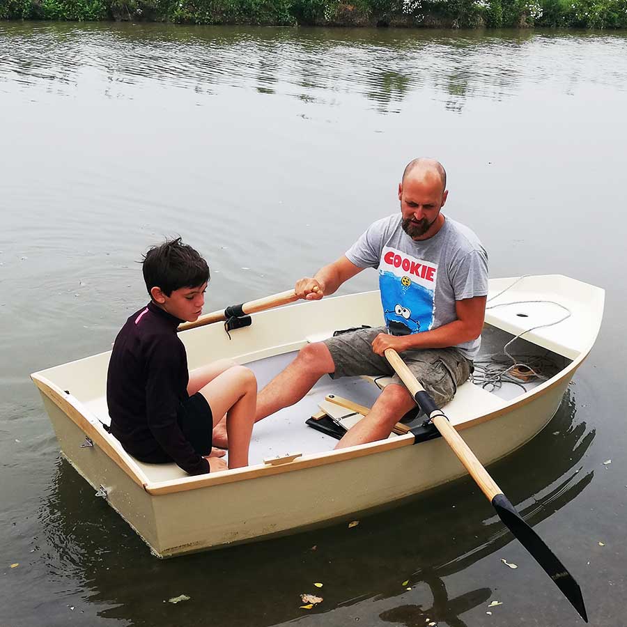 Homme et enfant à bord d’un petit bateau en bois Sardinette, en train de ramer sur un plan d’eau calme.
