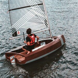 Voilier PRAM en bois naviguant sur l’eau, barré par un marin équipé d’un gilet de sauvetage.