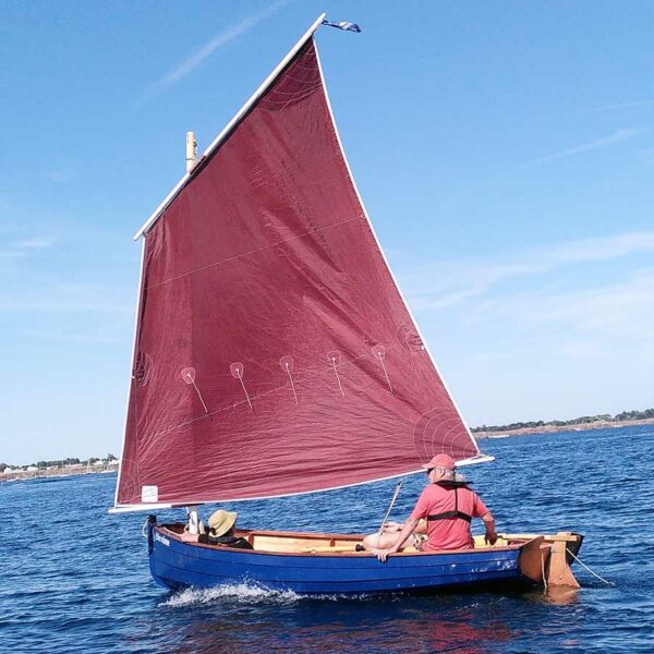 Voilier Jolie Mome bleu à voile bordeaux en navigation côtière avec trois personnes à bord